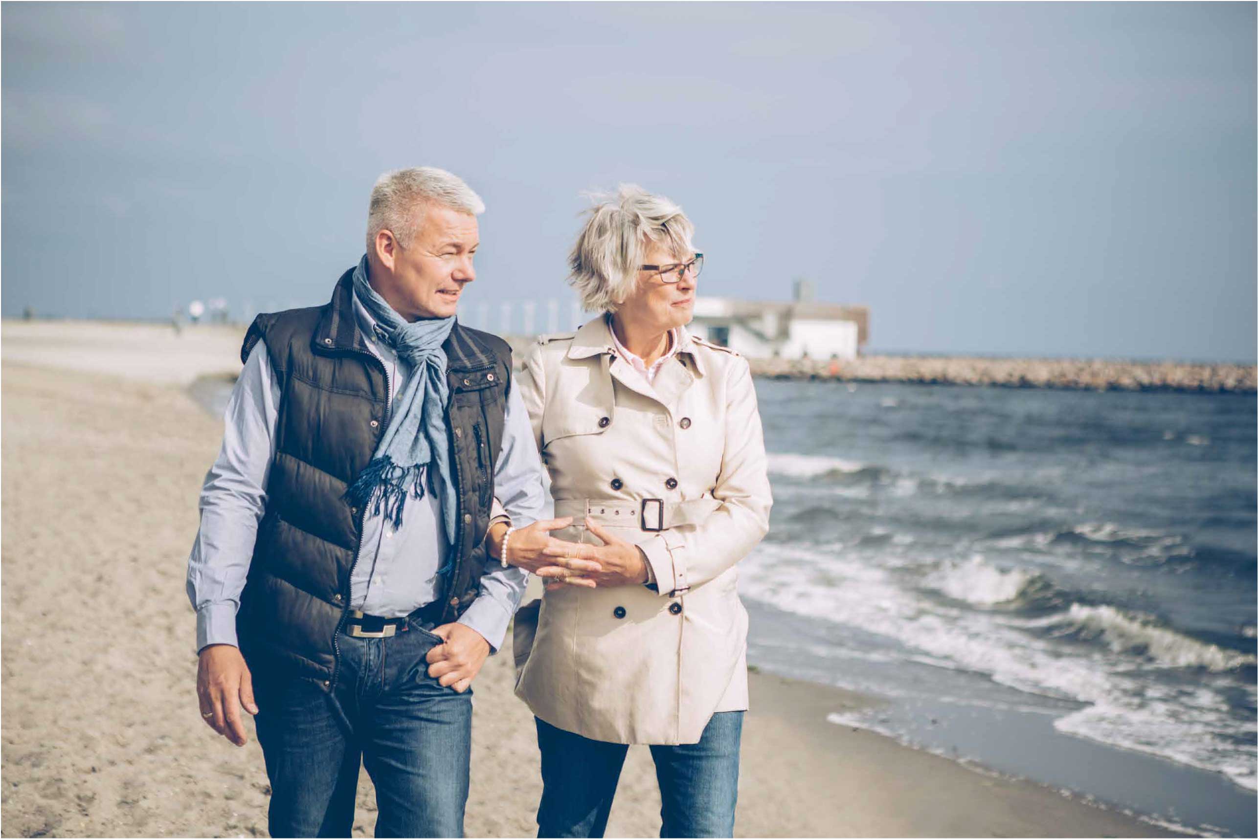 Modent ægtepar går tur på en strand med hinanden under armen.